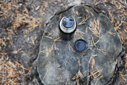 Black thermos bottle and a cup of tea standing on a stump surrounded by fallen pine tree needles, top view. Picnic in autumn park