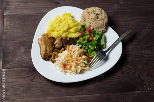 Dinner plate with fried fish, cabbage and vegetable salad, mashed potato, bun on brown wooden background