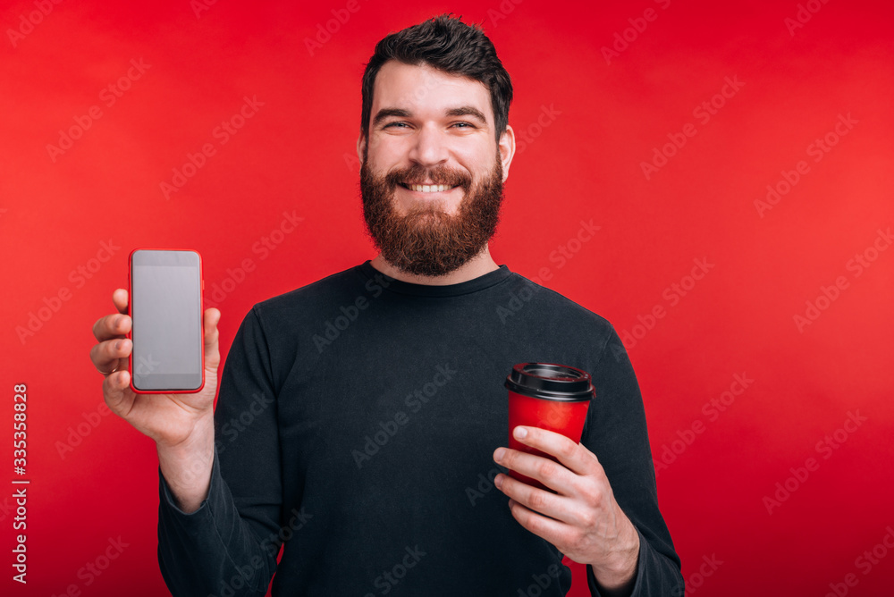 Portrait of smiling man with beard holding cup of coffee to go and showing smartphone screen Portrait of smiling man with beard holding cup of coffee to go and showing smartphone screen