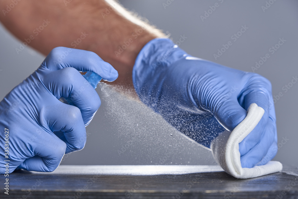 Hands in rubber gloves treat the table with a disinfectant spray on a grey background. Disinfection of premises - obrazy, fototapety, plakaty 