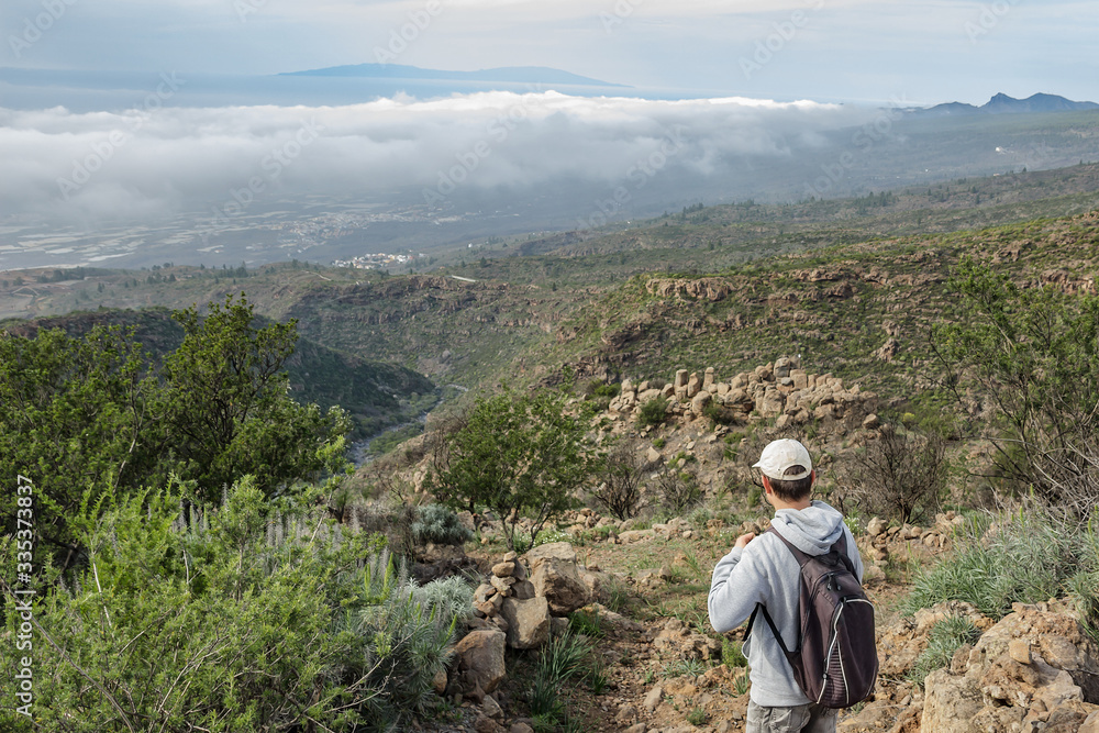 A young man in a cap with a backpack behind his back travels along a route in the Teide National Park. Hiking by the mountain trail surrounded by endemic vegetation and fields of lava rocks. Tenerife