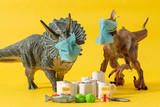 Two plastic dinosaurs with face masks and groceries on yellow background