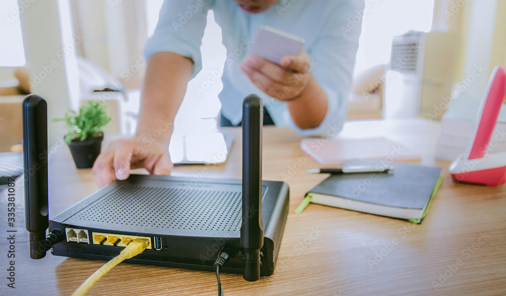Fototapeta premium closeup of a wireless router and a man using smartphone on living room at home ofiice, equipment for working from home, while in quarantine isolation during the Covid-19 health crisis.