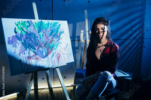 Girl artist sitting on a chair in a dark Studio near painted abstract acrylic painting.