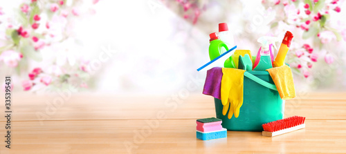 Bucket or basket with cleaning items on wooden table with spring flowers background. Washing set with copy space banner.