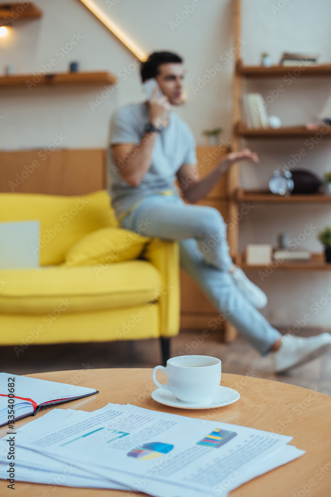 Selective focus of coffee table with papers, cup of coffee and notebook with freelancer talking on smartphone on sofa Selective focus of coffee table with papers, cup of coffee and notebook with freelancer talking on smartphone on sofa