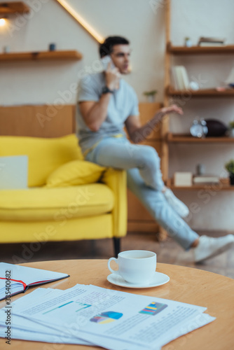 Selective focus of coffee table with papers, cup of coffee and notebook with freelancer talking on smartphone on sofa