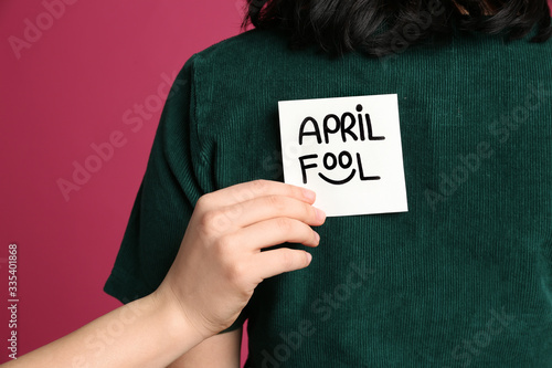 Woman sticking APRIL FOOL note to friend's back on pink background, closeup