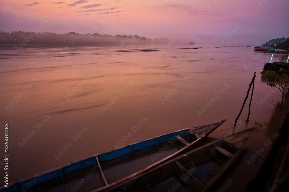 Beautiful, misty early morning sunrise over Rio Huallaga, Yurimaguas, in the Peruvian Amazon region. The pink, purple and orange light color the picture. Typical wooden fisher boats are in foreground.