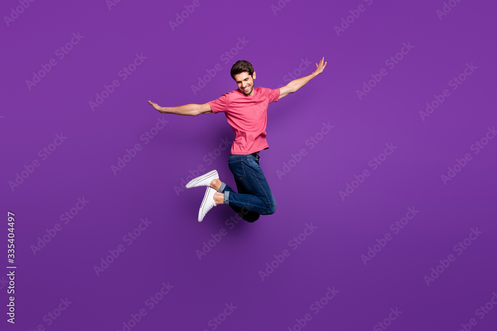 Full size profile photo of handsome attractive guy jump high up spread arms dance moves flight wear casual pink t-shirt jeans footwear isolated purple color background