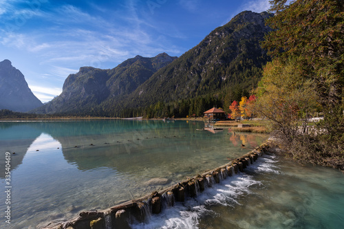Italian Dolomites, Lago di Dobbiaco - Toblacher See - calm blue-green water of lake in afternoon sun with shores covered with trees. On the right, water falls over a wooden weir.