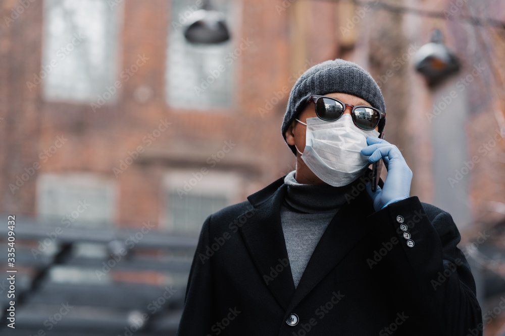 Sick man wears protective mask on face during virus pandemic, has telephone conversation with friend, wears black coat and shades, poses outdoor. Protect your health. Coronavirus, panic concept Sick man wears protective mask on face during virus pandemic, has telephone conversation with friend, wears black coat and shades, poses outdoor. Protect your health. Coronavirus, panic concept