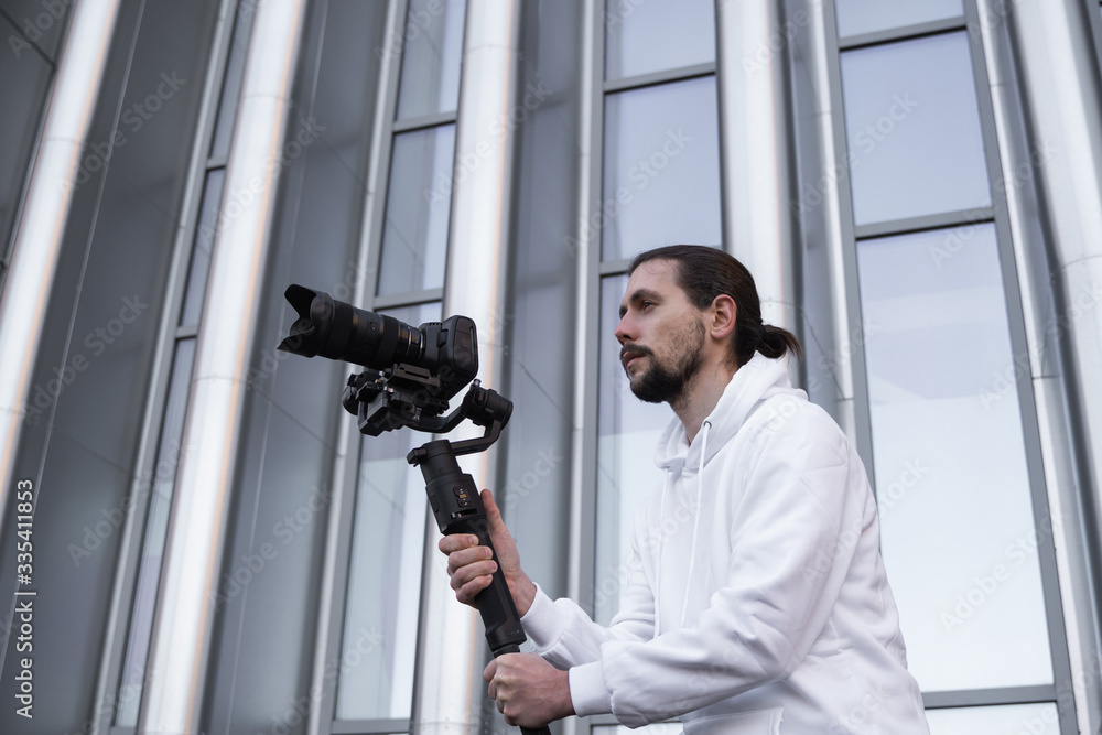 Young Professional videographer holding professional camera on 3-axis gimbal stabilizer. Pro equipment helps to make high quality video without shaking. Cameraman wearing white hoodie making a videos. Young Professional videographer holding professional camera on 3-axis gimbal stabilizer. Pro equipment helps to make high quality video without shaking. Cameraman wearing white hoodie making a videos.