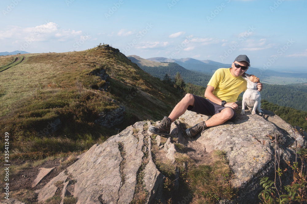 Alone tourist sitting on the edge of cliff with white dog against the backdrop of an incredible mountain landscape. Sunny day and blue sky