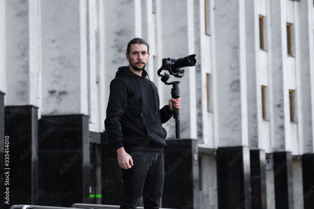 Young Professional videographer holding professional camera on 3-axis gimbal stabilizer. Pro equipment helps to make high quality video without shaking. Cameraman wearing black hoodie making a videos. Young Professional videographer holding professional camera on 3-axis gimbal stabilizer. Pro equipment helps to make high quality video without shaking. Cameraman wearing black hoodie making a videos.