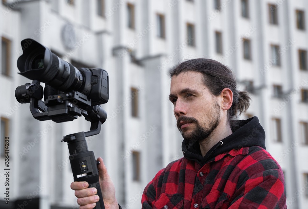 Young Professional videographer holding professional camera on 3-axis gimbal stabilizer. Pro equipment helps to make high quality video without shaking. Cameraman wearing red shirt making a videos. Young Professional videographer holding professional camera on 3-axis gimbal stabilizer. Pro equipment helps to make high quality video without shaking. Cameraman wearing red shirt making a videos.