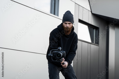 Bearded Professional videographer in black hoodie holding professional camera on 3-axis gimbal stabilizer. Filmmaker making a great video with a professional cinema camera. Cinematographer. photo