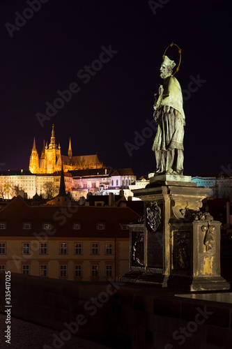 Jan Nepomucky, St. John of Nepomuk, the most famous Czech saint, his Statue on Prague Charles Bridge with the Prague gothic Castle in Night without People at the time of Coronavirus, Czech republic