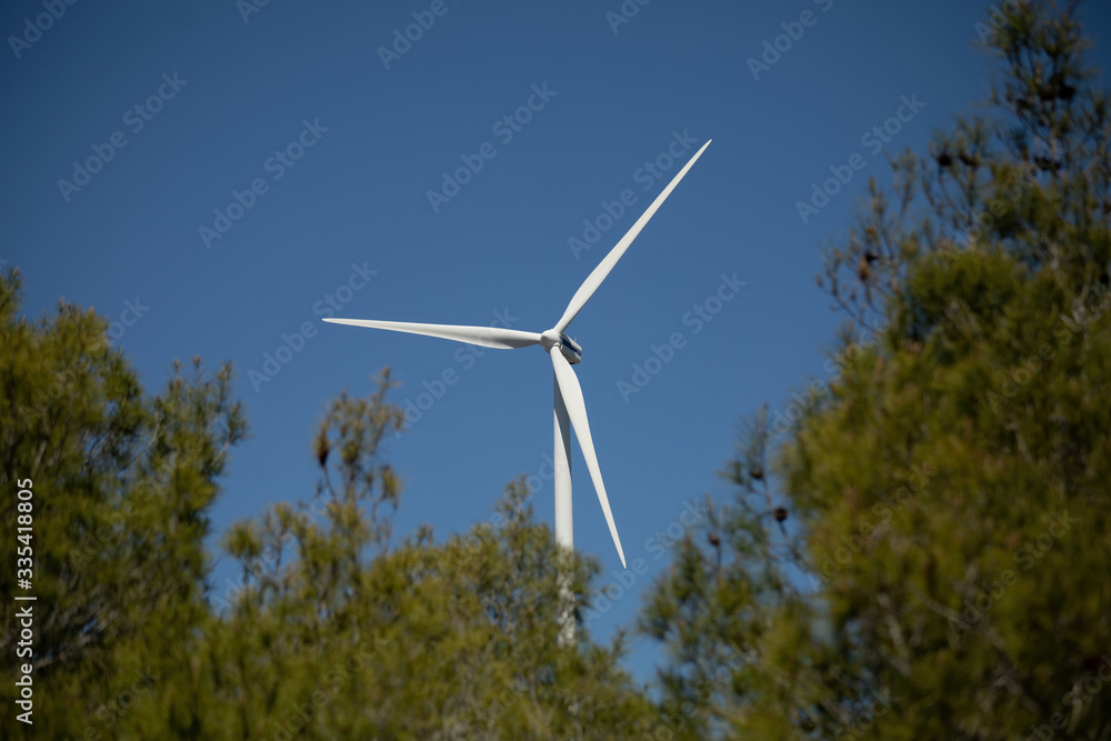 White wind turbines that generate electricity from renewable sources of nature surrounded by trees on a sunny day. Eco green energy. White wind turbines that generate electricity from renewable sources of nature surrounded by trees on a sunny day. Eco green energy.