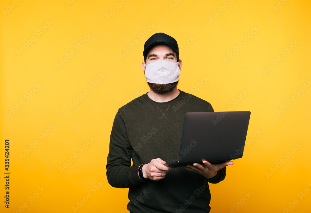 Portrait of young man with facial mask using laptop over yellow background