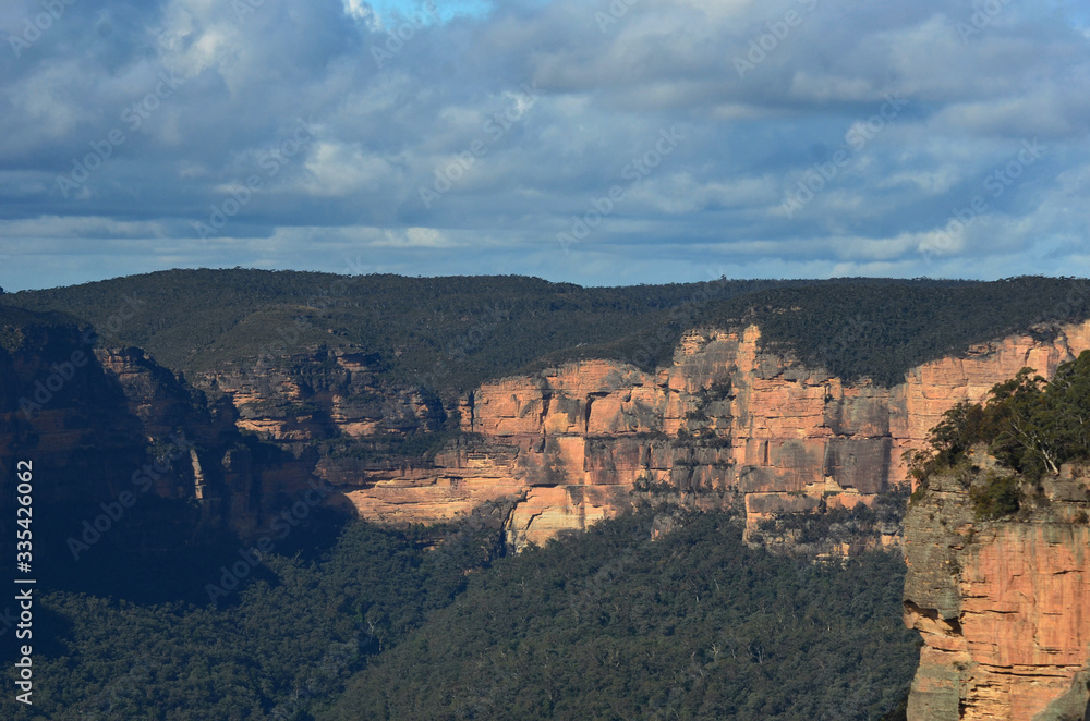 Sandstone cliffs are bathed in sunlight. They are surrounded by forest. The sky is overcast. They form part of the Megalong Valley in the Blue Mountains, Australia.