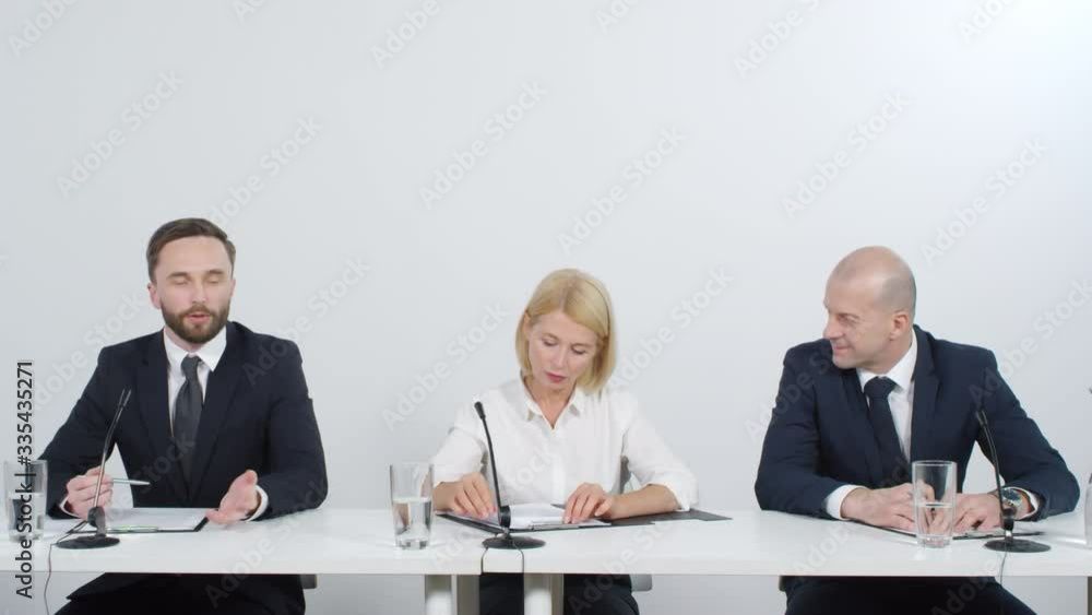Young male politician giving a conference talk while his colleagues discussing something at desk Young male politician giving a conference talk while his colleagues discussing something at desk