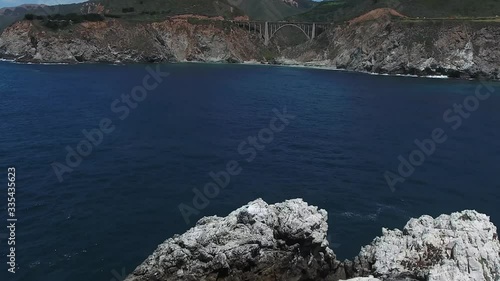 Aerial rising from behind big rock in blue water reveal Bixby Creek Bridge canyon cliffs and mountain side Big Sur