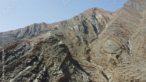 A stunning aerial pull back and tilt down revealing a lone cyclist riding on a narrow gravel road along a steep gorge, surrounded by stunning mountains makred with fascinating geological features.