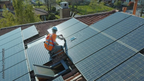 Aerial shot of technician with hard hat and safety equipment installing and working on maintenance of photovoltaic panel system installed on home domestic roof top, urban landscape. Wide angle camera.
