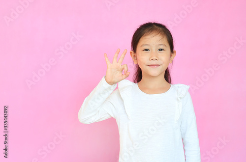 Beautiful asian little child girl making ok sign isolated over pink background. Portrait of smiling kid show confident hands symbol.