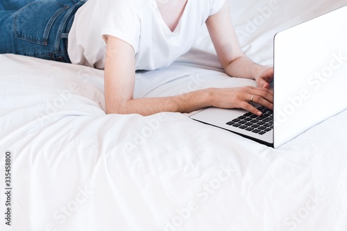 home office in a bed. young woman wearing white shirt and blue jeans and typing on a white and black laptop keyboard on white crumpled sheets