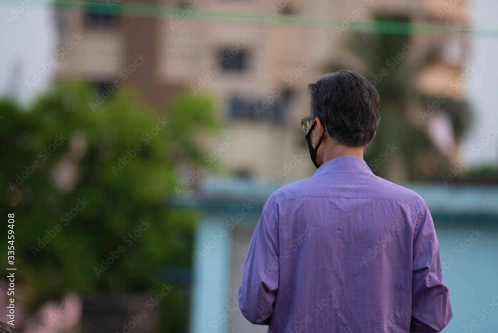Back portrait of an Indian old man with corona preventive mask and water bottle on a rooftop in home isolation.Indian lifestyle, disease and home quarantine. Back portrait of an Indian old man with corona preventive mask and water bottle on a rooftop in home isolation.Indian lifestyle, disease and home quarantine.