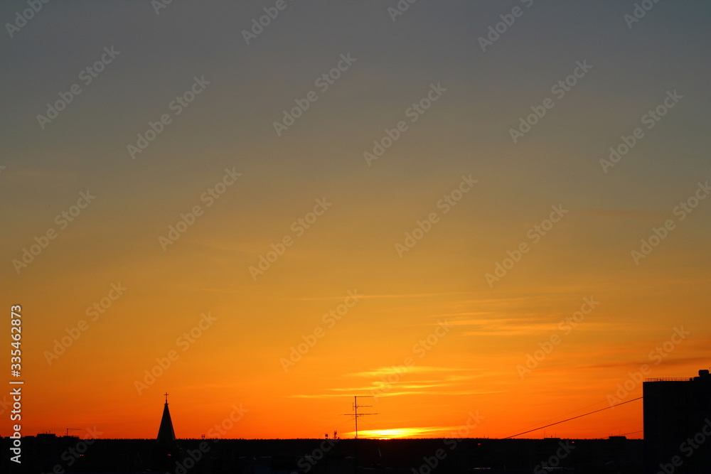 Naklejka premium Sun below the horizon and the roof of the church with a cross, television antenna, roof of a multi-storey urban building in the background in the background fiery dramatic orange sky at sunset or dawn