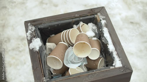 Close up garbage can full of paper cups. Someone throws another paper cup at him.