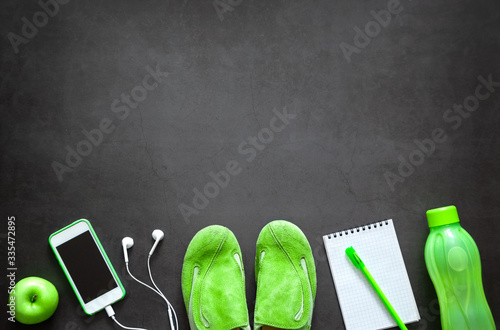 Green fitness equipment: sports shoes, a water bottle, an apple, a phone with headphones, a notebook with a pen. Healthy lifestyle. Concept. With copy space.