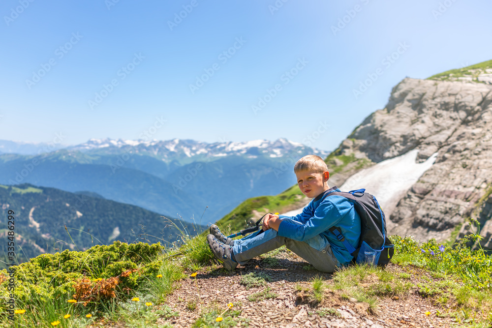 A boy traveler with trekking poles and a backpack is resting in the summer on top of a mountain with snow.