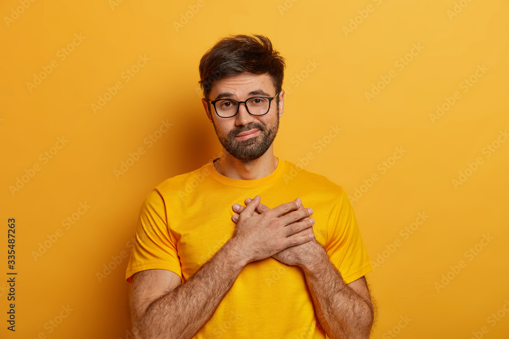Touched calm man presses hands to heart, being thankful to someone, expresses gratitude and good feelings, wears yellow t shirt and spectacles, hears congratulations after being promoted at work Touched calm man presses hands to heart, being thankful to someone, expresses gratitude and good feelings, wears yellow t shirt and spectacles, hears congratulations after being promoted at work