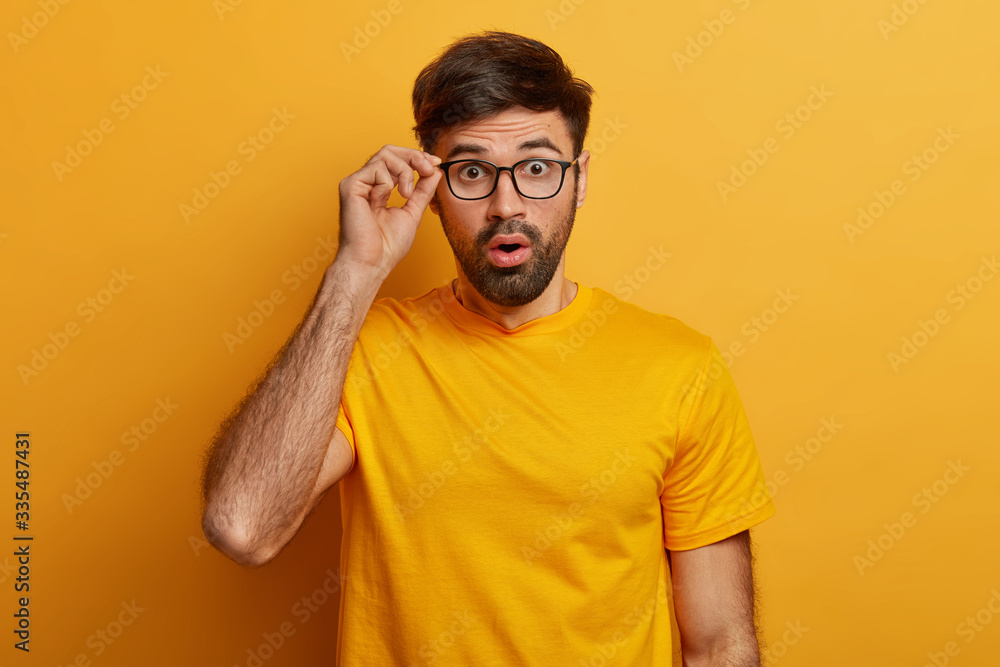 Man in stupor learns bad shocking news, keeps hand on rim of spectacles, has eyes popped out, cannot believe in fake relevation, dressed casually, isolated on yellow background, feels astounded