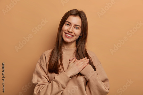 Touched pretty woman makes gratitude gesture, presses palms to heart, melts of adorable lovely gift received from friend, feels sympathy and passion, appreciates gift, stands against brown background