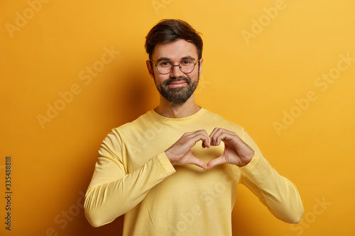 Canvastavla Handsome bearded adult man holds hands in shape of heart, expresses his affection to girlfriend, adores someone, says be my valentine, dressed in near sweater, isolated on yellow background