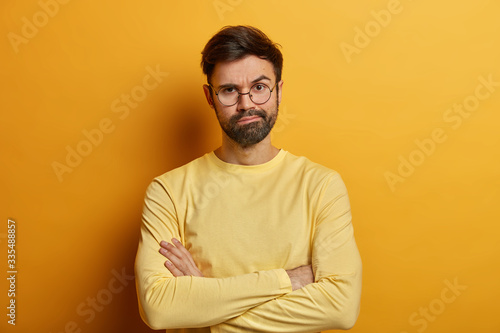 Serious man frowns face, stands impatient, keeps arms folded, waits for explanations, being jealous, raises eyebrow, wears transparent glasses and long sleeved sweater, isolated on yellow background