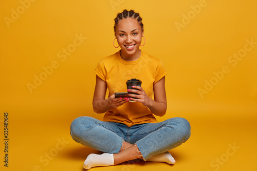 Relaxed young Afro American woman sits crossed legs in lotus pose, enjoys spare time and coffee break, holds disposable paper cup, scrolls internet social network, isolated on yellow background © wayhome.studio 