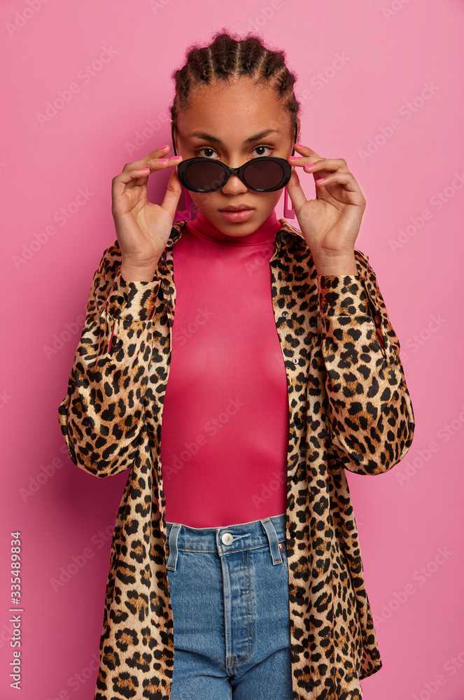 Naklejka premium Vertical shot of serious looking fashion model looks through sunglasses, wears leopard shirt and jeans, poses for magazine cover, isolated on pink background. People, style and beauty concept