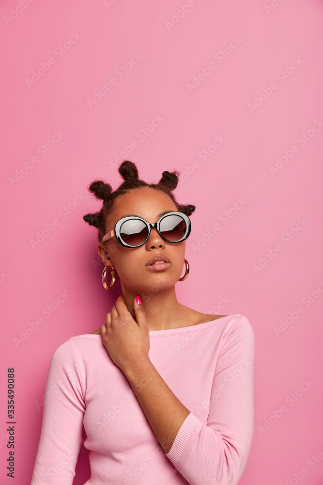 Glamour stylish woman keeps hand on neck, has confident look aside, wears sunglasses, earrings and fashionable jumper, has funny hairstyle with combed buns, poses against pink wall, copy space above