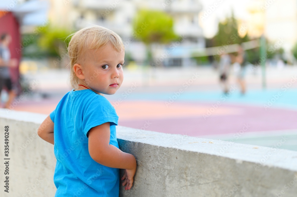 Naklejka premium a little three year old boy in a blue t-shirt watches teenagers playing with a ball on an outdoor sports field, dreaming of playing sports