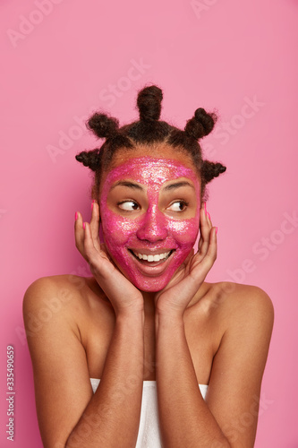 Photo of cheerful young woman touches cheeks gently, wants to have smooth glowing skin after applying beauty mask, has well groomed body, looks aside with smile, has combed knots, stands indoor