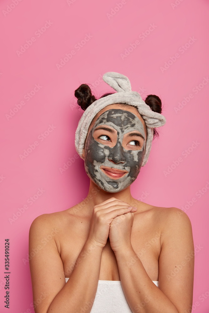 Natural beauty, skin care concept. Dreamy pleased Asian woman applies clay mask on face, keeps hands together under chin, looks gladfully aside, uses anti aging beauty product after taking shower