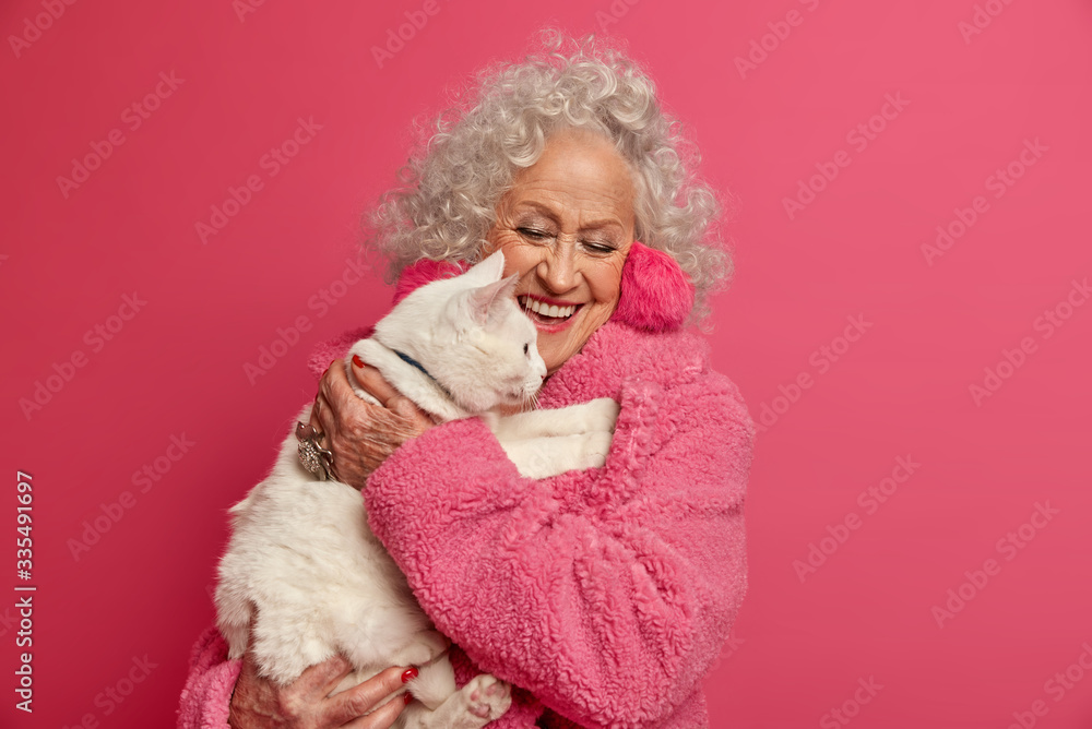 Old people stay at home during quarantine. Happy elderly curly woman dressed in domestic robe, holds white cat on hands, being on self isolation, feels not lonely in company of favorite pet. Old people stay at home during quarantine. Happy elderly curly woman dressed in domestic robe, holds white cat on hands, being on self isolation, feels not lonely in company of favorite pet.