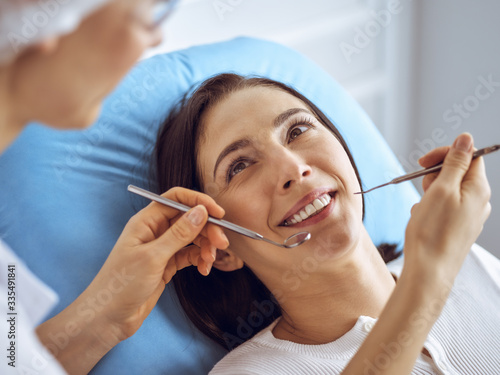 Smiling brunette woman being examined by dentist at dental clinic. Hands of a doctor holding dental instruments near patient s mouth. Healthy teeth and medicine concept