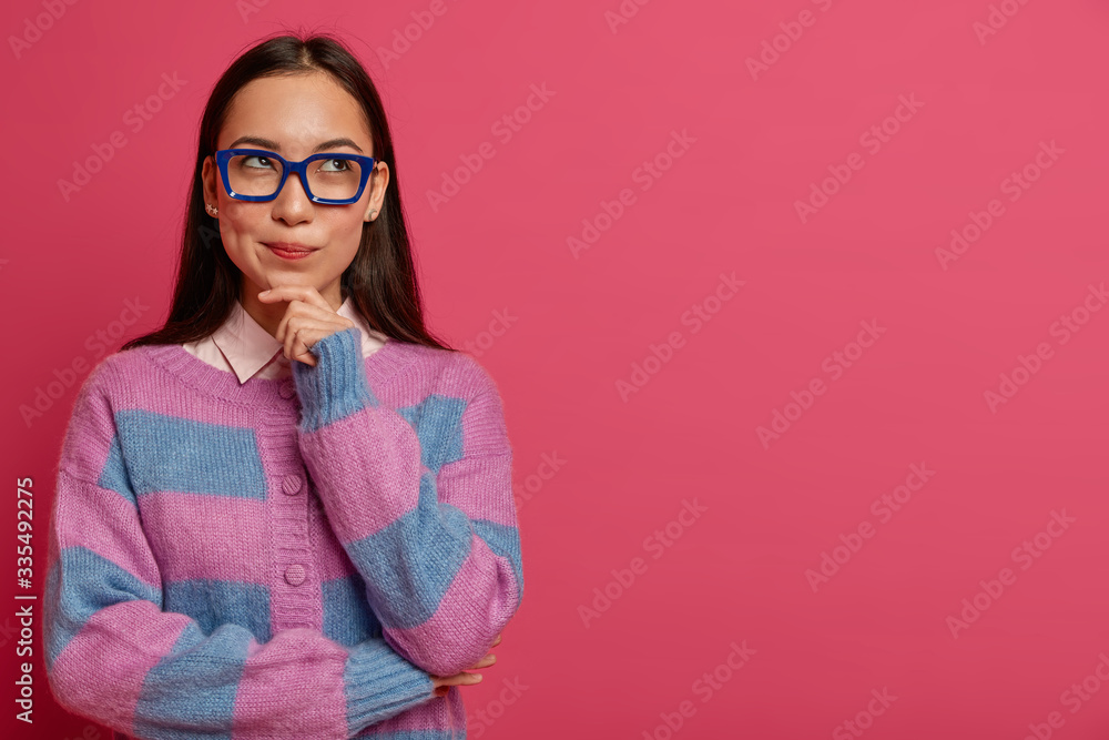 Thoughtful brunette female student wears big transparent glasses, looks to right side, holds chin, makes up great plan for future project, wears striped jumper, makes choice and faces decision